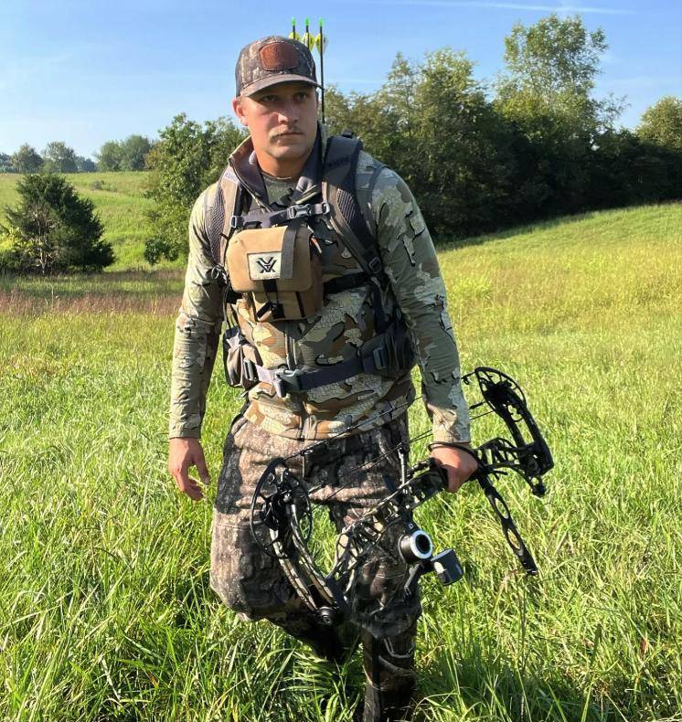 Nate in camo walking a hayfield with his compound bow
